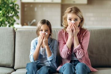 Sad caucasian mother and daughter with a cold are sitting at home on the sofa with napkins,...