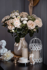 A creamy bouquet of roses and chrysanthemums, candles in the rays of light on a black table. The interior of the house is in the style of Provence. The concept of home comfort and decor in the house.