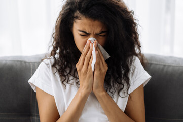 Ill african american young woman blowing running nose, she sneezing on a wipes, sit on sofa, sick...