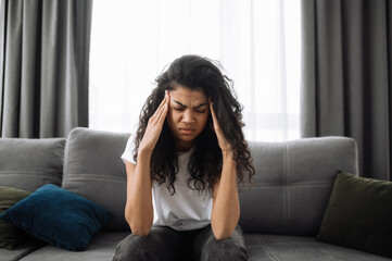 Photo of a sad african american young woman. Brunette bored at home, tired or stressed. Upset girl sitting on the sofa, having a headache or feeling depressed