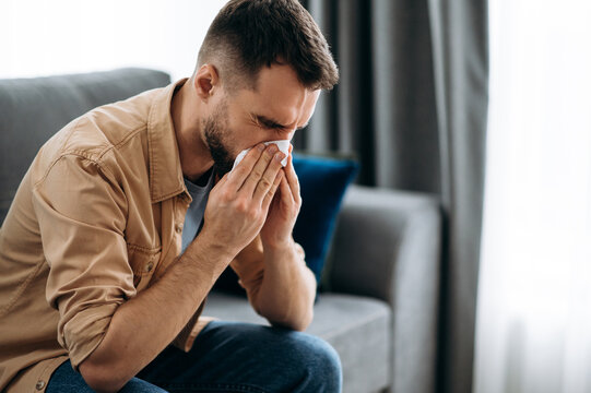 Sick caucasian guy sits on couch feel unhealthy, he suffering from rhinitis snuffles, having respiratory infection, get flue,  sitting at home at quarantine