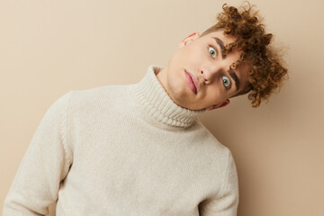 a close portrait of a handsome, funny man, with curly hair, standing on a beige background in a light turtleneck, looking pleasantly into the camera holding his head tilted
