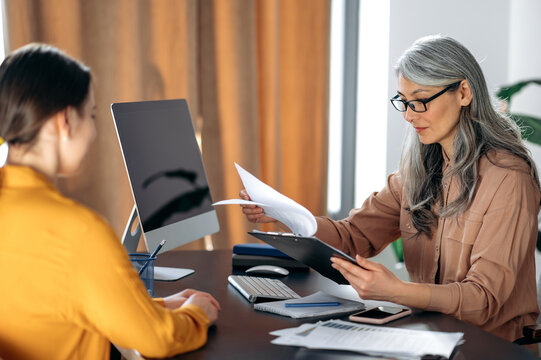 Friendly female mature hr manager listening and looks a resume of a new applicant introduction for a job. Gray-haired female employer talks and meets with female job seeker.