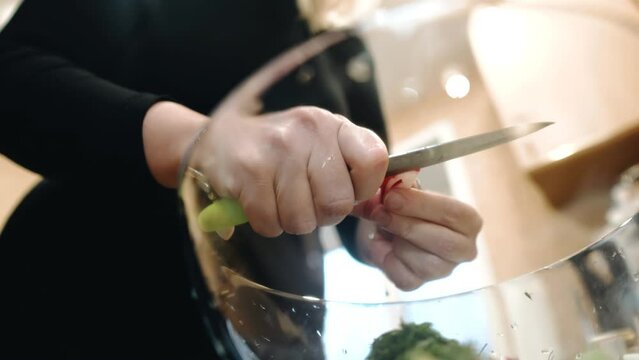 A woman cuts radishes into a transparent cup with a knife. She's making a salad for dinner