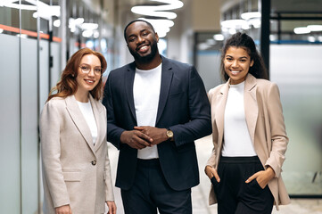 Confident successful colleagues stand in modern office. Portrait of multiethnic smiling business people in formal wear. Friendly male and female employees look at the camera, teamwork concept
