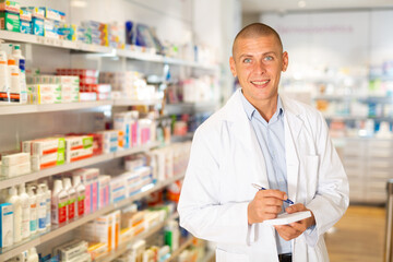Portrait of an male pharmacist working in pharmacy during the pandemic, standing in trading floor and makes important notes