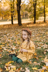 Fototapeta na wymiar A beautiful little blonde girl in a yellow dress sits in an autumn park holding maple leaves.
