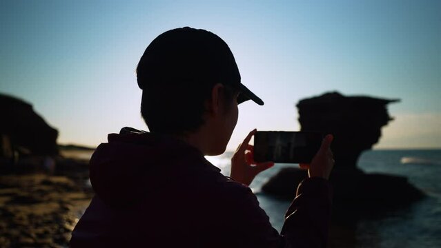 Over the shoulder of a woman taking a photo of Teapot Rock with her phone, Prince Edward Island at sunset, creating beautiful lens flare.