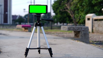 Blank green screen mobile smartphone taking live photo or video on tripod on blurred nature background in social technology concept.  Mockup design