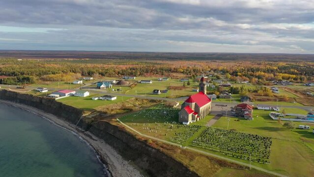 Drone footage over Chaleur Bay towards a graveyard, near eroding cliffs and over the colorful red roof of the Church of Saint Simon and Saint Jude.