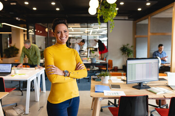 Portrait of smiling young biracial businesswoman standing with arms crossed in creative office