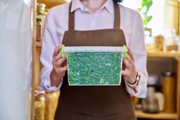 Woman with container of frozen green onions, in kitchen in pantry near freezer.