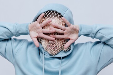 horizontal studio photo of a strange, tortured man in a light blue hoodie covering his face with...