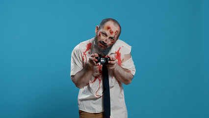 Spooky bizarre looking zombie with modern gaming controller playing games on console. Sinister mindless brain-eating monster playing with electronic wireless gamepad on blue background.