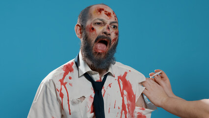 Growling scary zombie getting vaccine imunisation shot on blue background. Frightening brain-eating creepy monster with deep and bloody scars and wounds getting flu shot injection. Studio shot © DC Studio