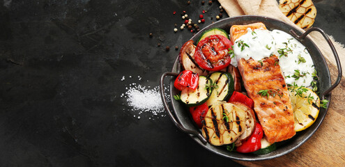 Salmon steak with vegetables and white sauce on dark background.