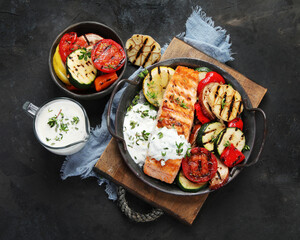 Salmon steak with vegetables and white sauce on dark background.