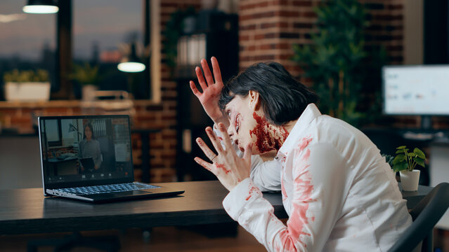 Creepy angry zombie in online video conference with asian colleague. Brain-eating walking dead corpse with bloody wounds getting irritated becuase of internet video call with company office coworker.