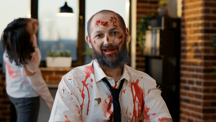 Obraz premium Mindless dumb looking zombie in office workspace sitting at table. Dangerous creepy brain-eating monster with deep and bloody scars and wounds looking at camera while sitting inside company workspace.