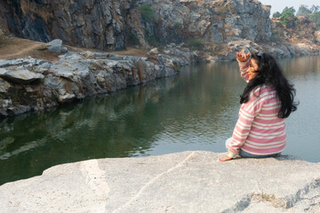 An Indian young woman sitting on rock by the lake at sunset or sunrise looking at view and enjoying the serene atmosphere. Girl relaxing on winter contemplating nature.