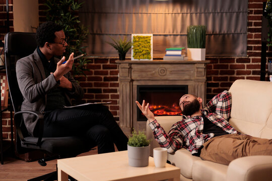 African american male therapist consulting man laying on couch, doing psychoanalysis to solve problems at therapy session. Talking about behavior issues and giving counseling advice.