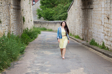 Lifestyle portrait of young stylish woman with long brunette hair walking on the street in old town, wearing yellow dress and denim jacket