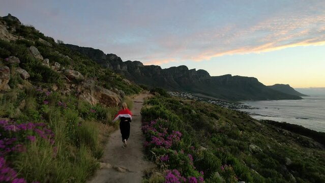 young blonde girl in red jacket running Lions Head Trail at sunset towards 12 Apostles in Cape Town, aerial