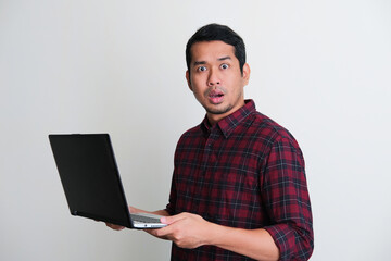 Adult Asian man looking camera with worried expression while holding his laptop