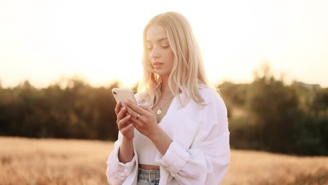 Young beautiful brunette woman in white shirt texting on her phone in the gold field during the sunset.