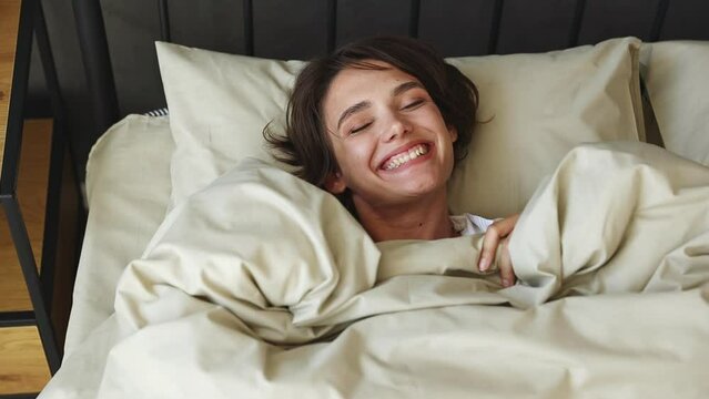 Young fun woman wears casual white shirt she lying in bed rest relax spend time in bedroom lounge home in own room hotel wake up crawled got out from under covers good mood day. Lazy morning concept