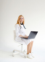 a doctor girl with red hair is working on a laptop sitting on a chair on a white isolated background.