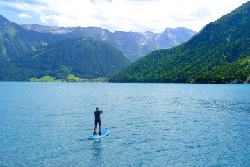 man sailing on sapboard, lake Achensee in Austria, green mountains rises above calm expanse of...