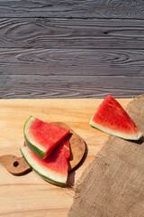 Ripe pieces of red cut watermelon with pits lie on a board, on a light yellow wooden table, against the background of a blue textured wooden wall.