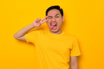Cheerful young Asian man in casual t-shirt showing peace sign with fingers over face isolated on yellow background. People lifestyle concept