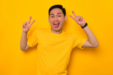 Fototapeta premium Cheerful young Asian man in casual t-shirt showing peace sign with fingers isolated on yellow background. People lifestyle concept