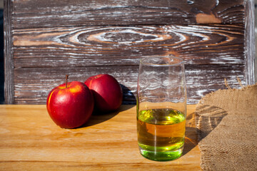 A glass of freshly squeezed apple juice and a red ripe apples on a table covered with a cloth against a wooden textured wall.