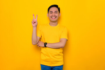 Cheerful young Asian man in casual t-shirt showing peace sign with fingers isolated on yellow background. People lifestyle concept