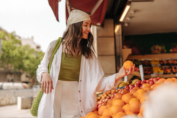 Beautiful young caucasian girl buys citrus fruits at outdoor market. Brunette wears casual clothes,...
