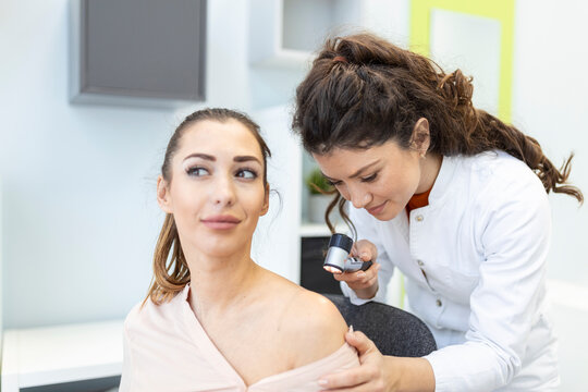 Dermatologist in latex gloves holding dermatoscope while examining attractive patient with skin disease. Female dermatologist examining patient with dermascope, looking for signs of skin cancer.