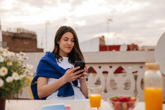 Cute young caucasian girl uses her smartphone sitting at juice table on terrace in morning. Brunette girl wears casual clothes in spring. Lifestyle concept