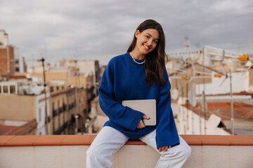 Smiling young caucasian woman holding tablet, looking at camera sitting on rooftop with beautiful view. Brunette wears casual blue sweater, jeans. Sincere emotions lifestyle concept