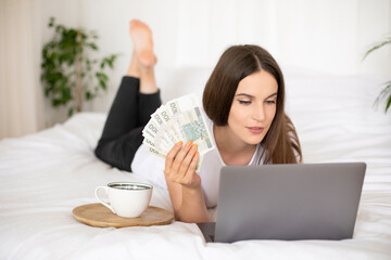 Young girl lying on a bed and holding polish zloty, uses laptop computer. Work from home