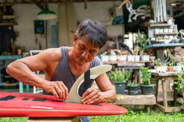 Asian senior man attaching fins on bottom of surfboard at home. Elderly male surfer enjoy outdoor lifestyle surfing at the sea on summer vacation. Retirement people activity and water sport concept