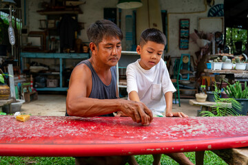 Happy Asian family grandfather teaching grandchild boy waxing surfboard surface. Senior man and...