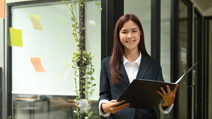 Young asian female manager in formalwear holding folder and standing near glass window in office