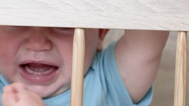 cute little baby boy is crying in the crib, holding the wooden bars with hands and pushing with feet. toddler legs through crib bars.nervous agitated kid. sunny morning