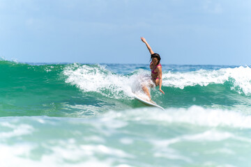 Asian woman surfer surfing and riding surfboard  the wave in the sea at tropical beach in sunny day. Healthy female enjoy outdoor activity lifestyle and water sport exercise surfing on summer vacation