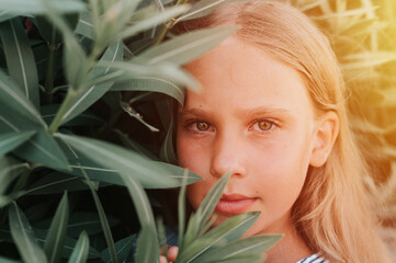 Naklejka premium portrait face of candid happy little kid girl of eight years old with long blond hair and green eyes on background of green plants during a summer vacation travel. gen z mental health concept. flare