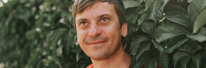 portrait of face happy smiling unshaven mature 40+ years man solo traveler with stubble and brown eyes on the background of green trees plants in nature on sunny summer day on vacation travel. banner