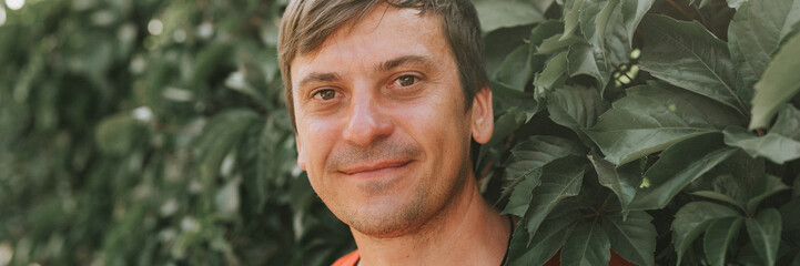 portrait of face happy smiling unshaven mature 40+ years man solo traveler with stubble and brown eyes on the background of green trees plants in nature on sunny summer day on vacation travel. banner